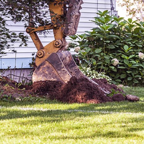 A large construction equipment excavator with a heavy, hinged, hydraulic, iron and steel excavation scoop shovel is digging, lifting and spilling the first load of dirt and rocks from the initial hole in the ground in this front yard lawn at the edge of a house exterior wall at the very beginning of a suburban residential district home improvement new in-law apartment home addition construction project.